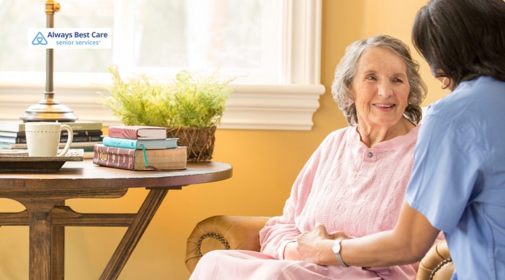 A caregiver in a light blue shirt smiles as she provides comfort and assistance to an elderly woman in a pink sweater, seated on a chair in a warm, cozy indoor setting.