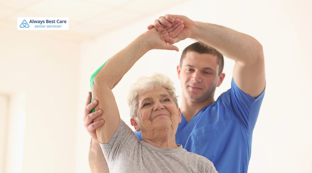A caregiver in blue scrubs helps a smiling senior woman stretch her arm during a gentle rehabilitation exercise.