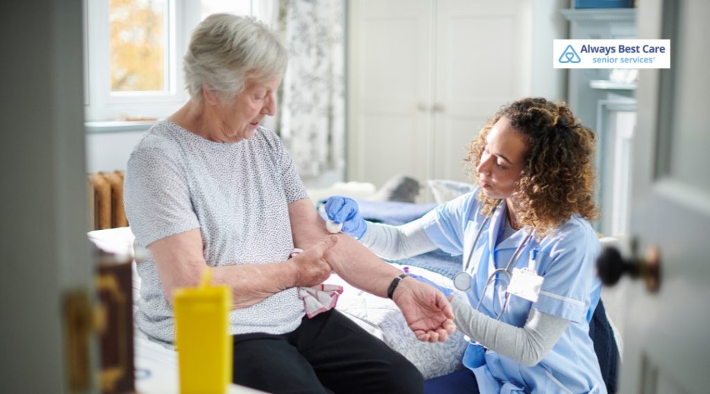 A nurse in blue scrubs provides in-home care by applying a bandage to an elderly woman’s arm as she sits on a bed.
