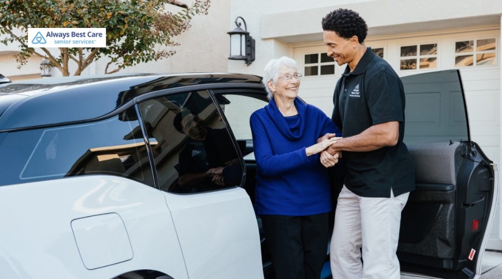 A caregiver warmly supports an elderly woman as she steps out of a car in front of a home.