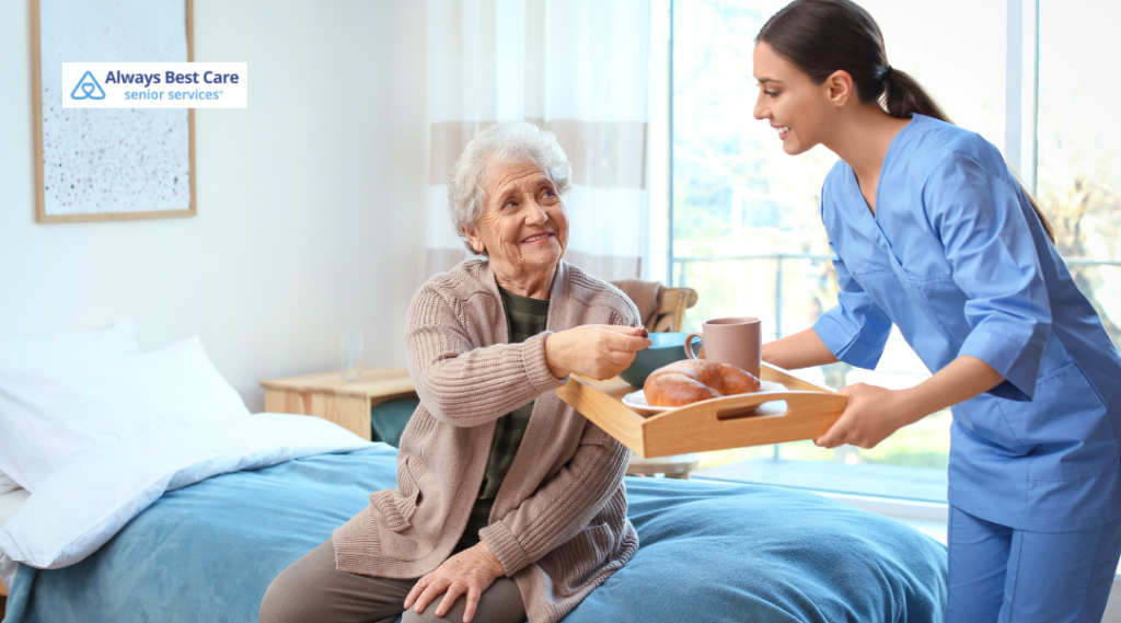 Caregiver serving breakfast to a smiling older woman seated on her bed.