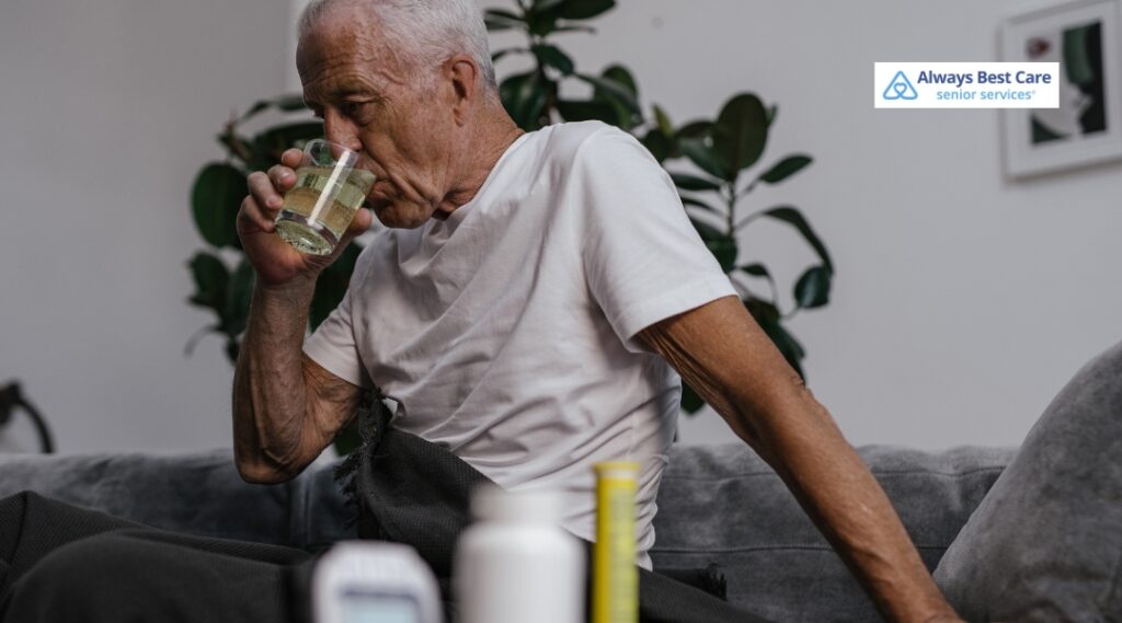 Older man drinking water while seated near medication containers at home.