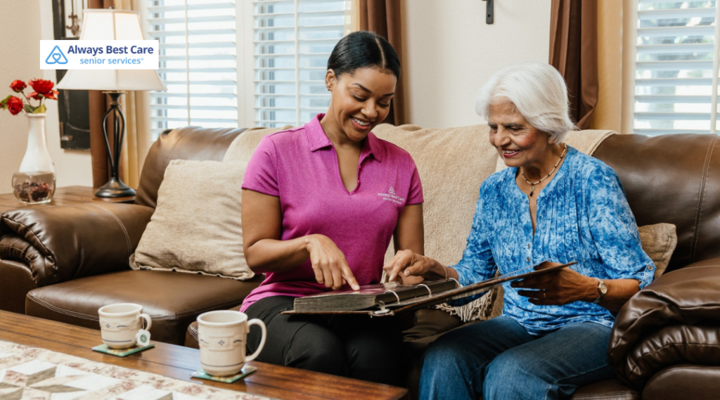 Caregiver and older woman smiling as they look through a photo album together at home.