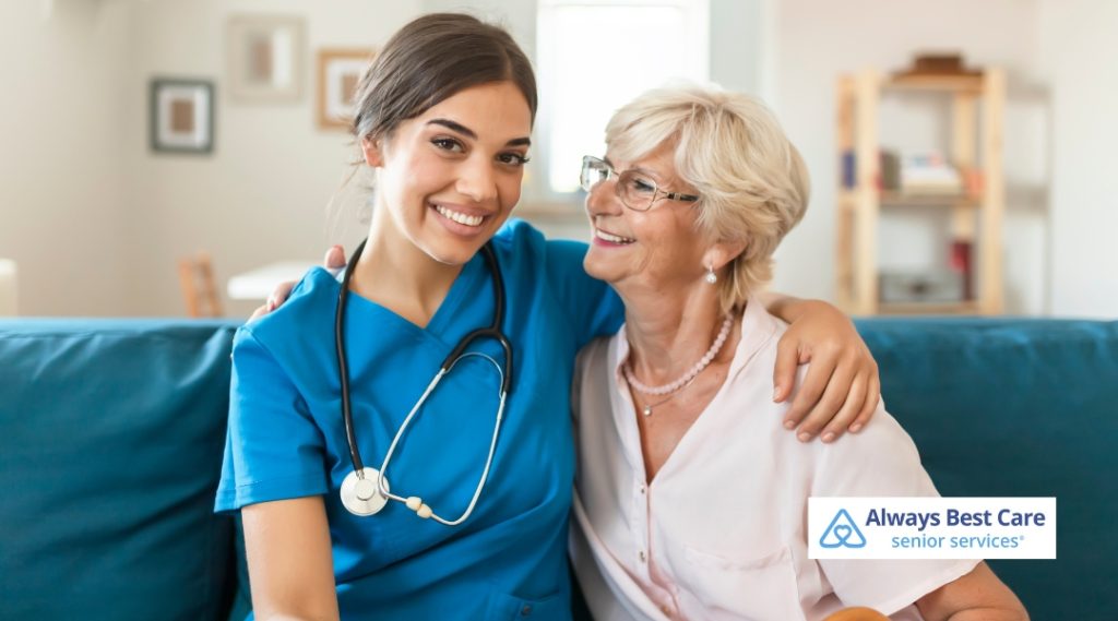 A senior woman and her caregiver are sitting together on a couch, smiling happily. The caregiver, wearing a blue uniform with a stethoscope, sits next to the senior. The Always Best Care logo is visible in the bottom right corner.