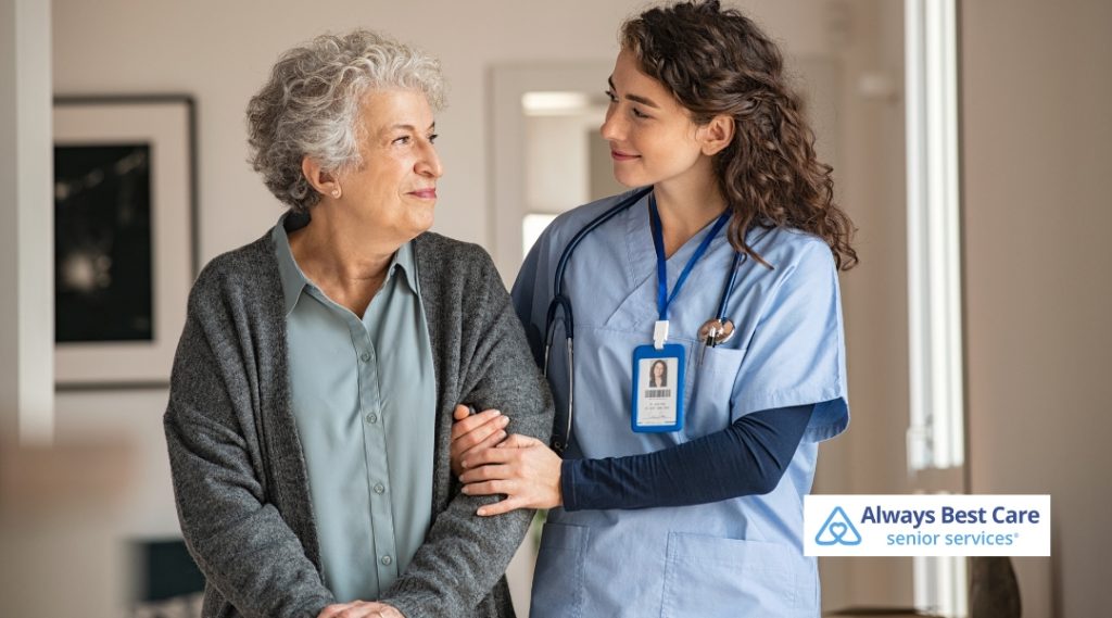 A caregiver is helping a senior woman walk while supporting her by holding her arm. The caregiver wears a blue uniform, and the image features the Always Best Care logo in the corner.