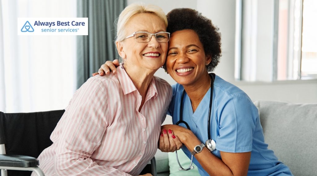 A senior woman and her caregiver smile at the camera, sitting close together. The caregiver is wearing a blue uniform, and the senior woman is in a pink shirt. The Always Best Care logo is displayed in the corner.