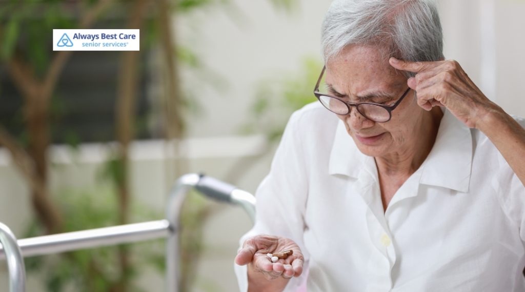 An older woman with short gray hair looks confused while holding medication in her hand and pointing to her temple. A walker is visible beside her, and the Always Best Care logo is in the top left corner.
