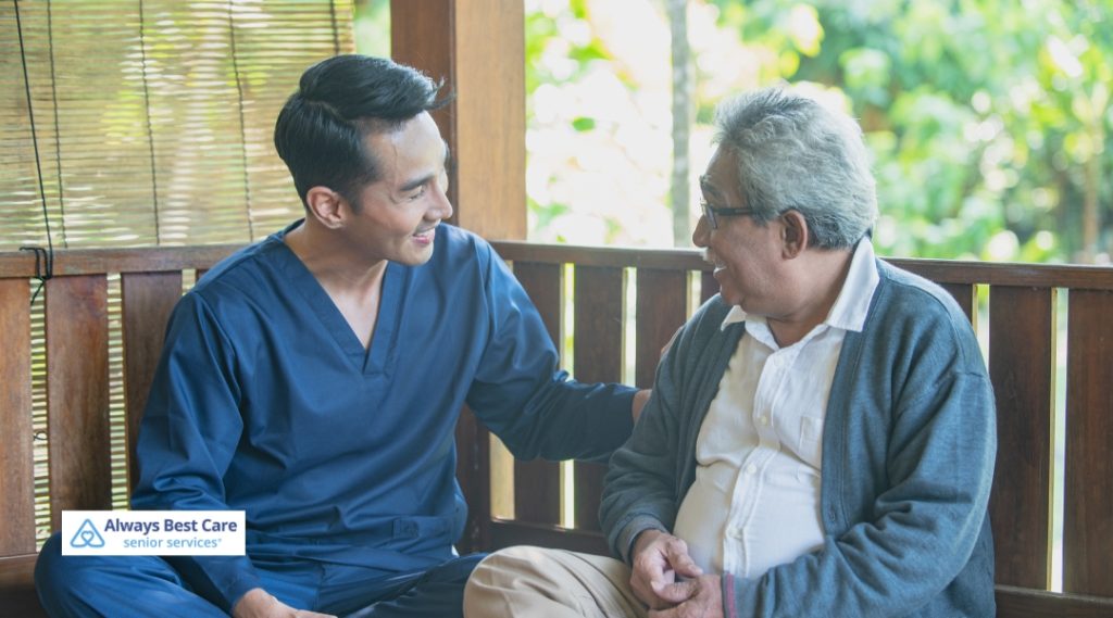 A male caregiver in dark blue scrubs shares a moment of connection with an elderly man, both smiling as they sit together on a wooden bench in a warm outdoor setting.