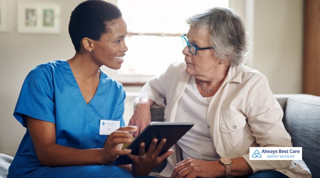 A female caregiver in blue scrubs holds a tablet while having a discussion with an elderly woman in glasses, both smiling and seated together in a bright, comfortable indoor space.