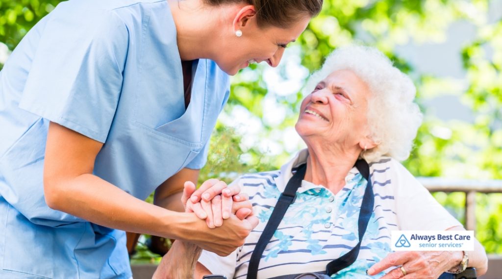 A caregiver in light blue scrubs holds the hands of an elderly woman in a wheelchair, both sharing a joyful, uplifting moment in a sunny outdoor setting.