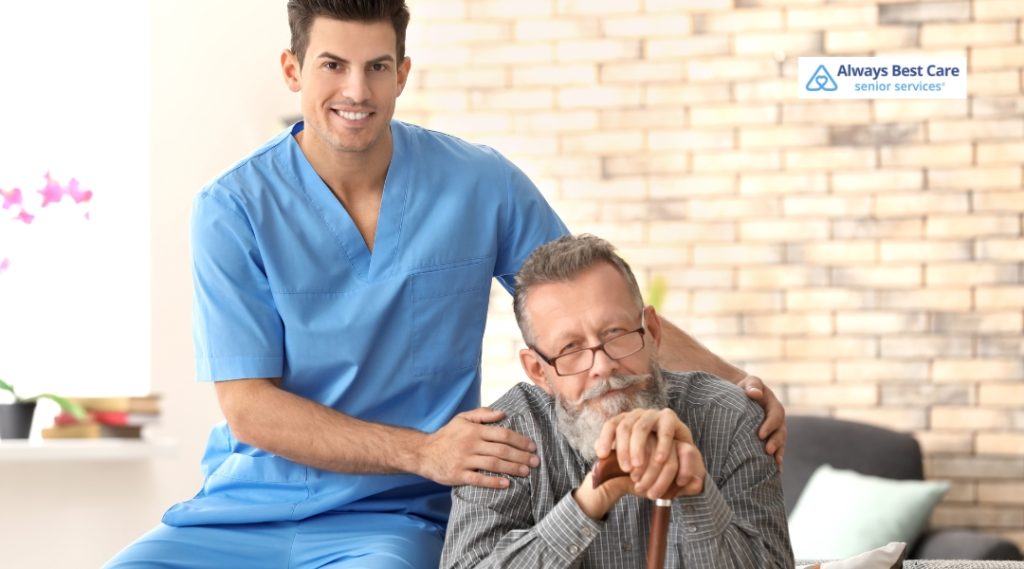 A compassionate caregiver in blue scrubs smiling beside an elderly man with a cane, offering a supportive presence in a homely, light-filled space.