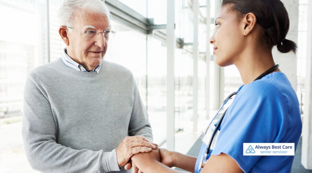 A caregiver in blue scrubs holding hands with an elderly man, offering a steady and caring presence in a modern and spacious indoor setting.