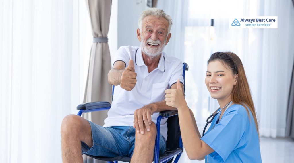 An elderly man and caregiver smiling and giving a thumbs-up, enjoying a positive moment together, with light-filled windows creating a bright, welcoming atmosphere.