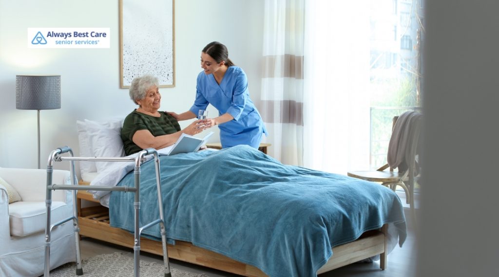 A smiling caregiver hands a glass of water to a senior woman sitting up in bed, with a walker nearby.