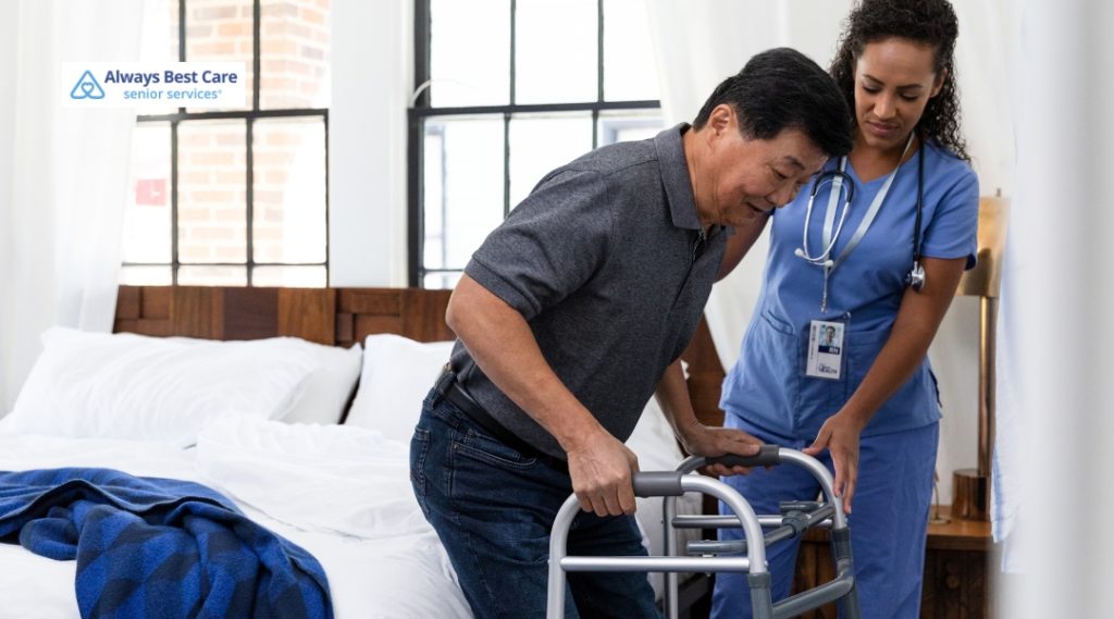 A nurse helps a senior man using a walker to stand up safely beside his bed.