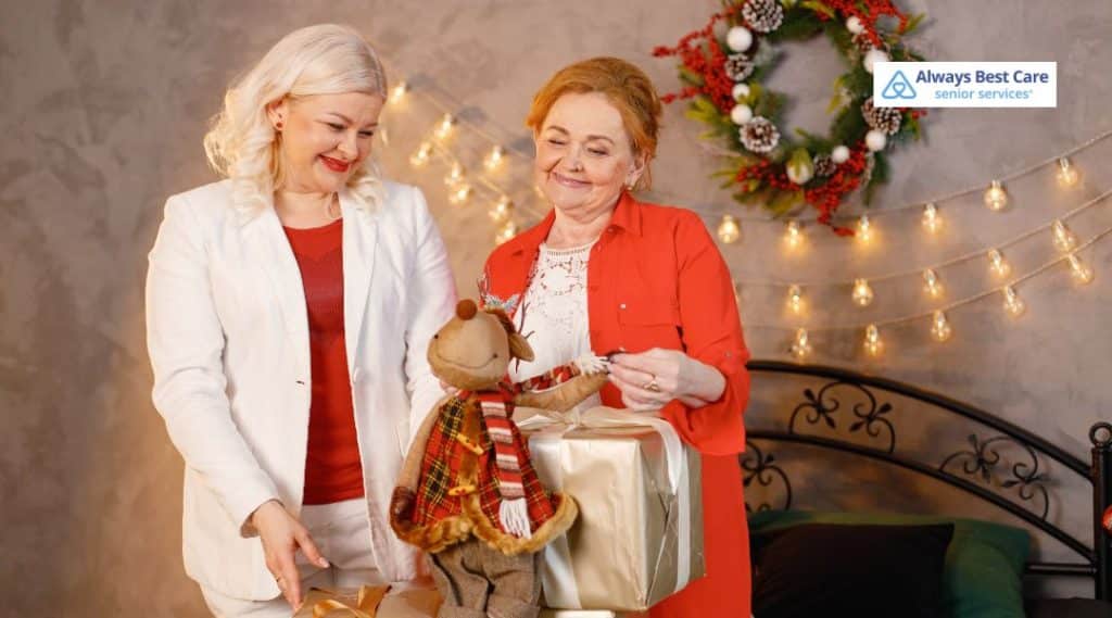 Caregiver and older woman smiling while opening a holiday gift together in a warmly decorated home, representing companionship and senior support from Always Best Care.
