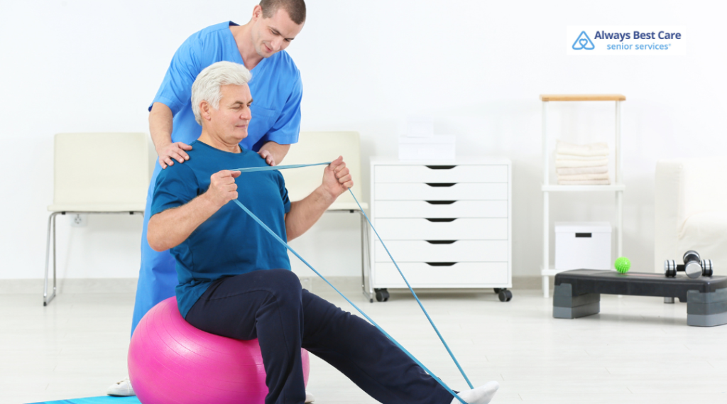 Senior man sitting on an exercise ball, stretching a resistance band while a caregiver in blue scrubs supports his shoulders.