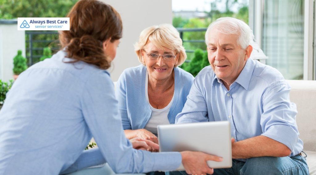 A female caregiver showing a tablet screen to an elderly couple sitting on a couch, both smiling and attentively looking at the device. The setting appears bright and comfortable, with a green outdoor background visible through the window.