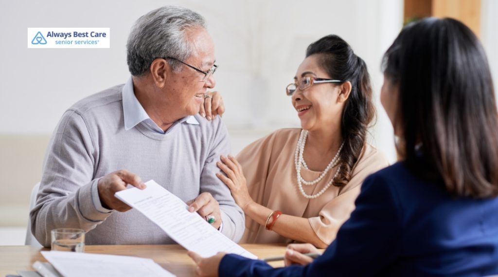 An elderly man and woman sitting indoors at a table, sharing a joyful moment while reviewing paperwork with a female caregiver. The couple looks happy and engaged, and the caregiver is handing them documents.
