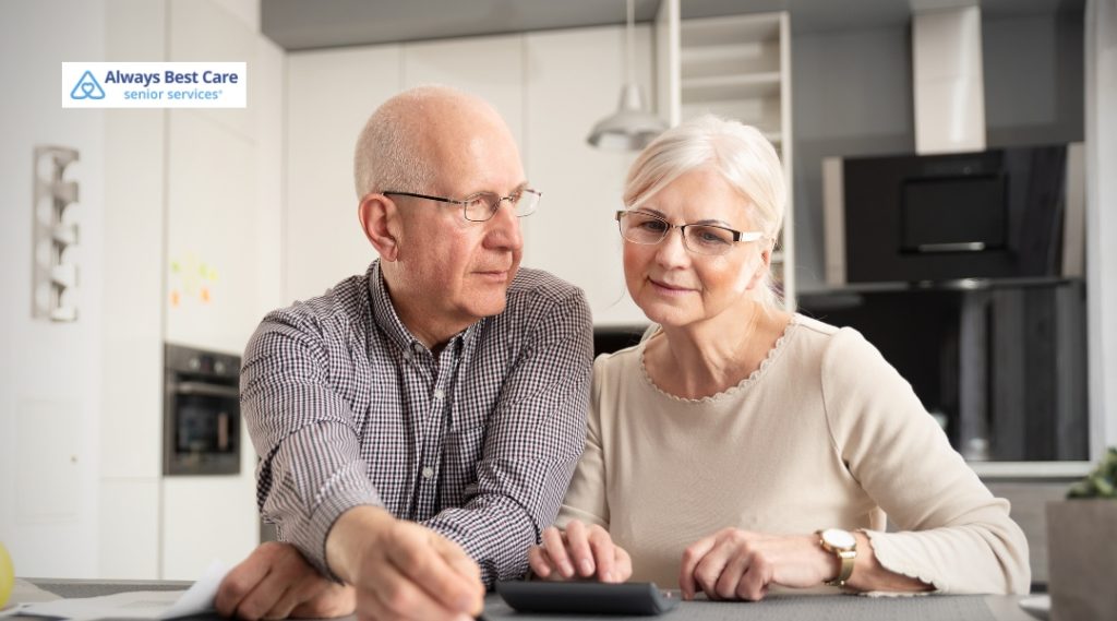 An elderly couple sitting close together at a kitchen table, focused on calculating or reviewing bills and paperwork, conveying a sense of partnership and mutual support in managing finances or care.