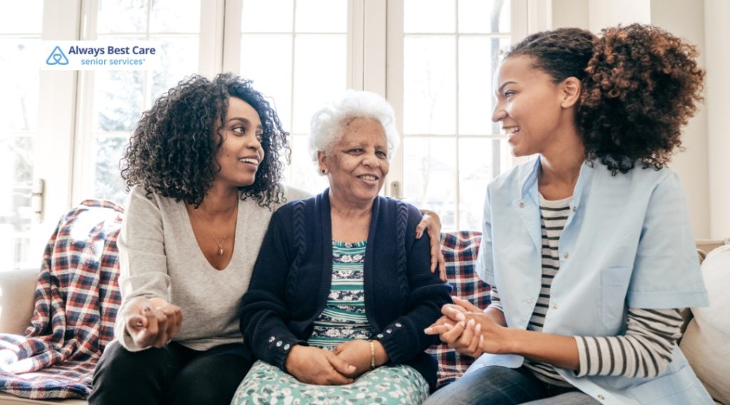 Two younger women sitting with an elderly woman in a cozy living room. The elderly woman is smiling warmly as the women engage with her, providing companionship and care in a bright, welcoming home environment.