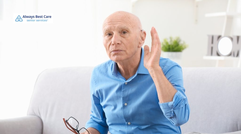 A senior man is sitting on a couch, raising his hand with a questioning expression, as if having trouble hearing or understanding.