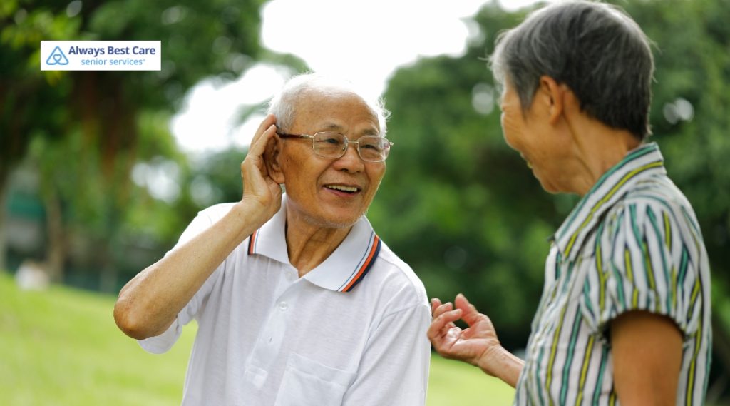 Two seniors, one of whom is holding his ear to better hear, appear engaged in a conversation outdoors in a natural setting.