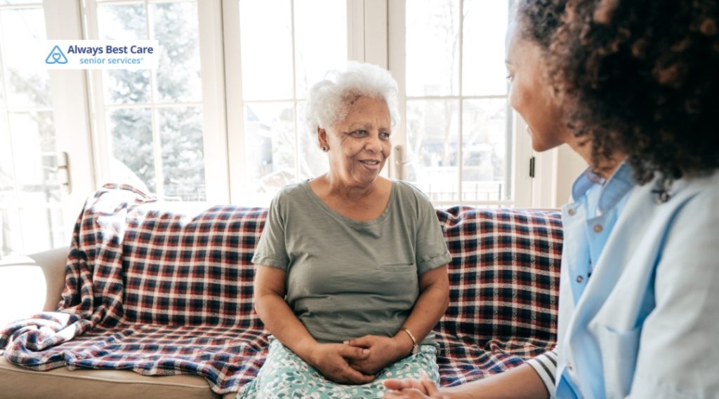 A senior woman is sitting on a couch with a caregiver, enjoying a conversation in a comfortable home setting.