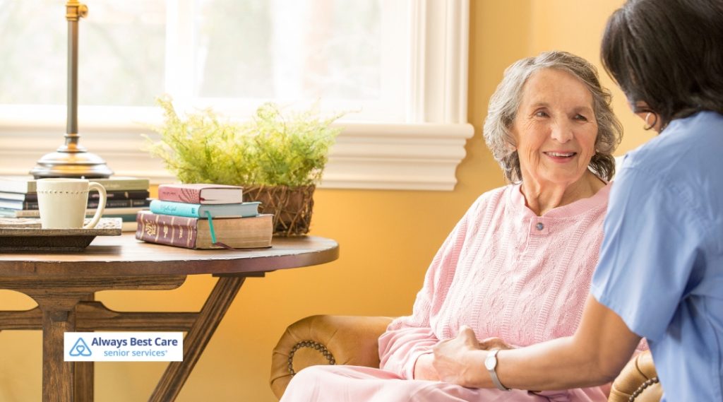 A female caregiver offers comforting support to an elderly woman sitting in an armchair, holding her hands. The woman looks content and reassured. A table with books and a cup is visible in the background. The Always Best Care logo is in the bottom left corner.
