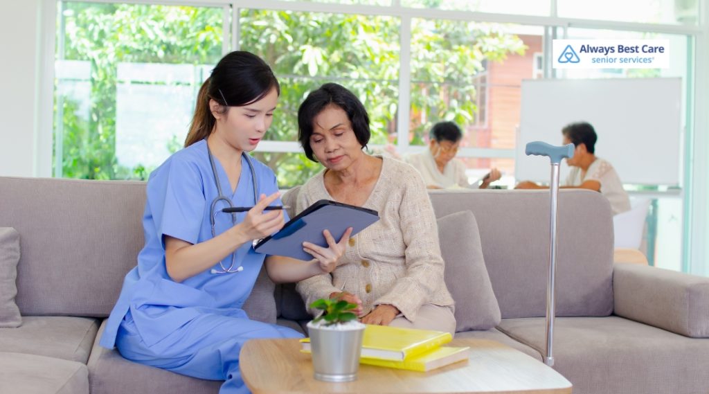 A caregiver in a blue uniform and a senior woman sit together on a couch, with the caregiver showing her something on a tablet, reflecting the personalized in-home care services provided by Always Best Care.