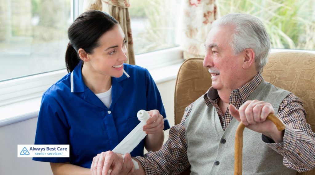 A caregiver in a blue uniform wraps a bandage around a senior man's hand while smiling at him, demonstrating the hands-on, attentive care Always Best Care provides to its clients.