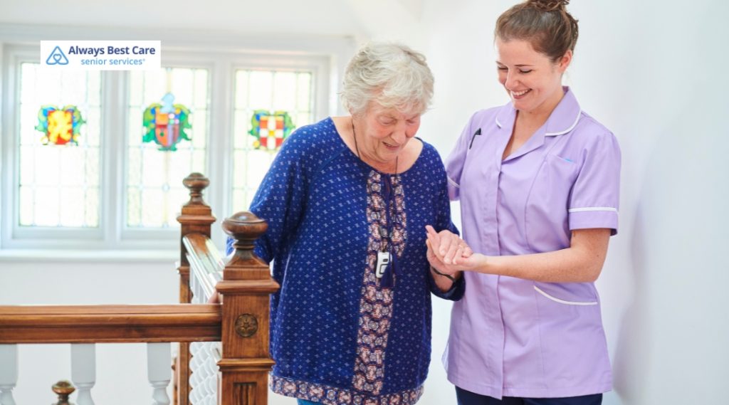 A caregiver in a purple uniform supports a senior woman while she walks down a staircase, highlighting the assistance Always Best Care offers for mobility and safety at home.
