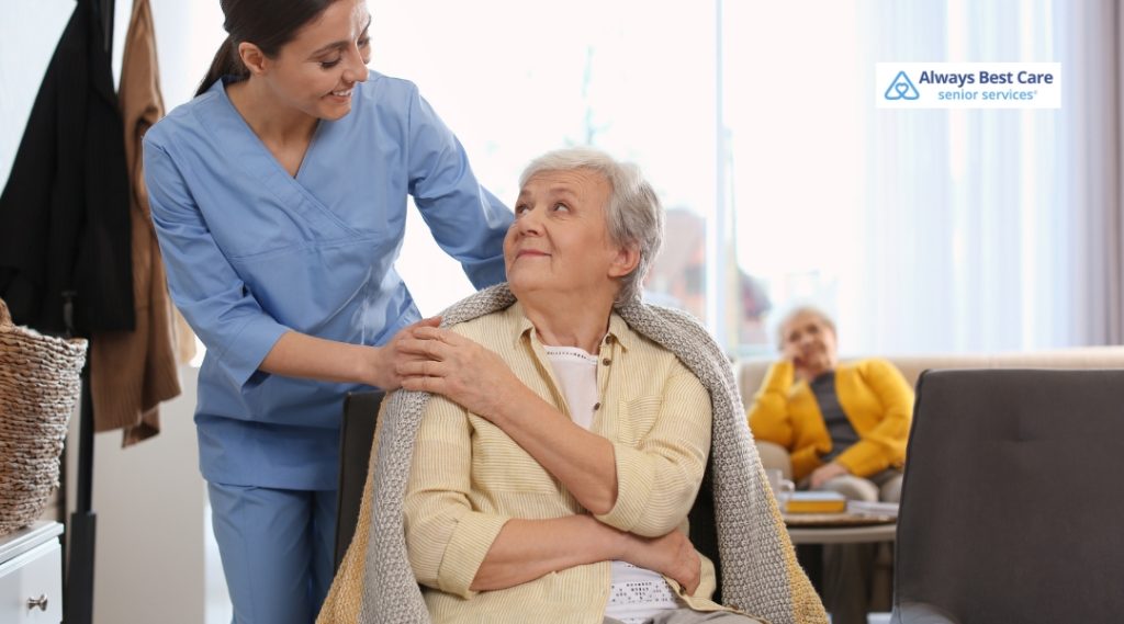 An elderly woman smiles warmly at a female caregiver draping a blanket over her shoulders, while another senior woman sits comfortably in the background.