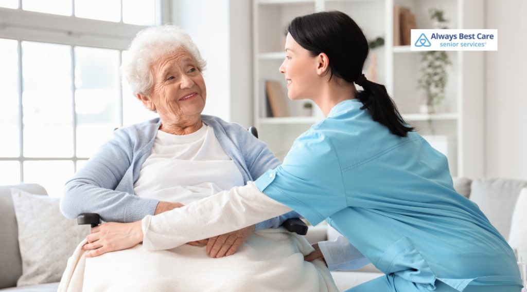 A smiling female caregiver in blue scrubs crouches to talk to an elderly woman sitting in a wheelchair, sharing a comforting and positive moment.