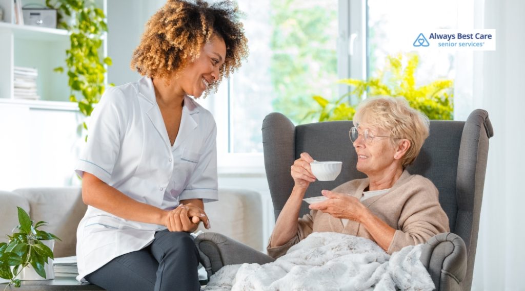 A cheerful caregiver chats with a senior woman enjoying a cup of tea while wrapped in a cozy blanket, seated in a bright and peaceful living room.