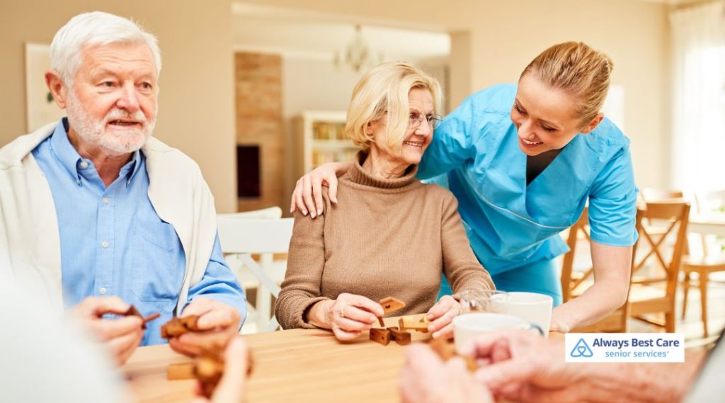 A group of seniors sits around a table engaging in wooden puzzle activities with a caregiver in blue scrubs offering gentle encouragement, fostering cognitive stimulation and social interaction in a bright, supportive care setting.