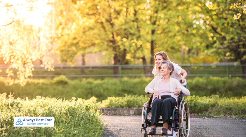 A caregiver gently pushes an elderly woman in a wheelchair along a sunlit outdoor path surrounded by greenery in a serene park setting. 