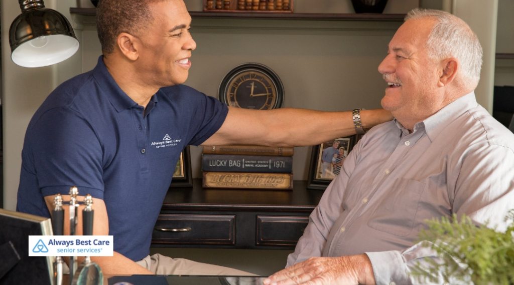 A caregiver in a blue shirt is smiling and providing support to an elderly man sitting at a table, showing care and friendly interaction in a cozy indoor setting.