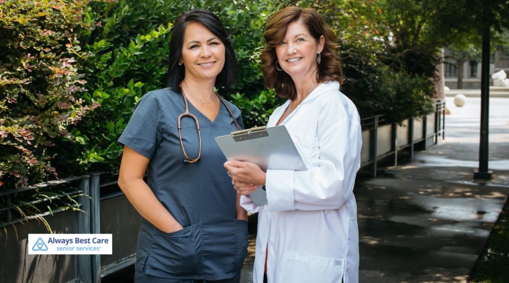 Two healthcare professionals, one in scrubs and the other in a white lab coat, stand outside in a garden, smiling and looking friendly, as they discuss care services together.