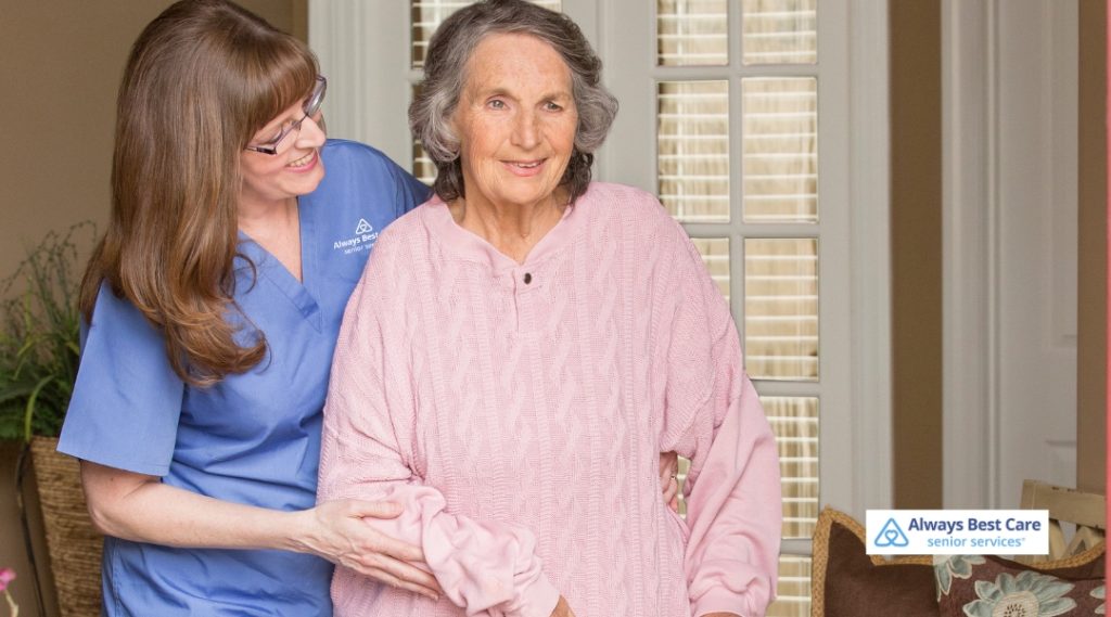 A caregiver in a blue shirt is helping an elderly woman in a pink sweater stand and walk with support, smiling as they assist each other in a cozy, welcoming home environment.