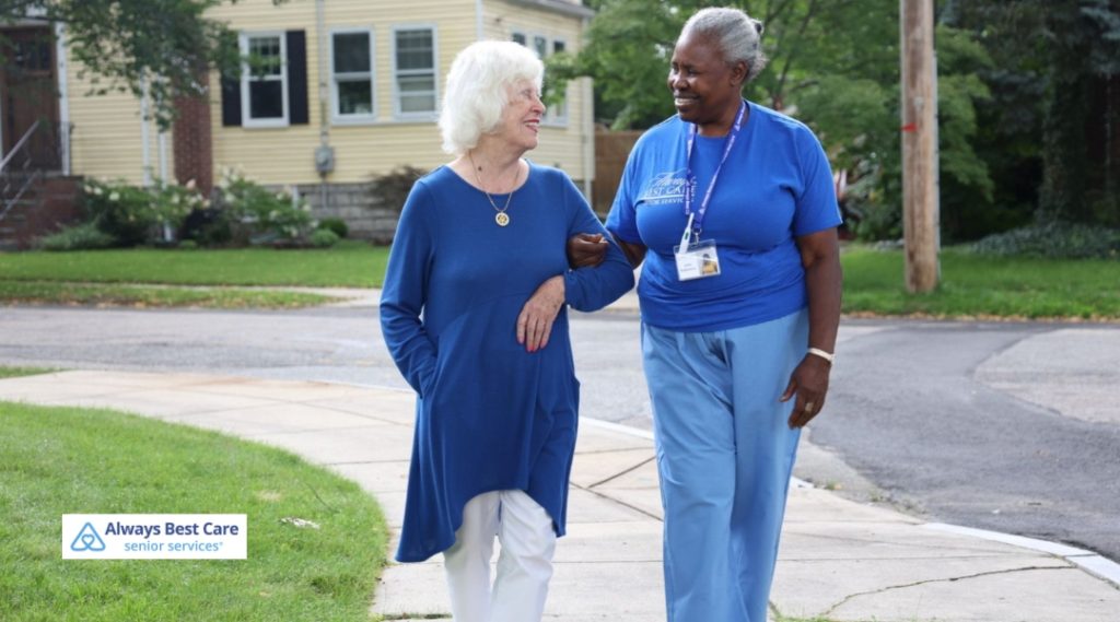 A caregiver and an elderly woman in a blue dress are walking outside together on a sunny sidewalk, with both smiling and enjoying the fresh air in a suburban neighborhood.