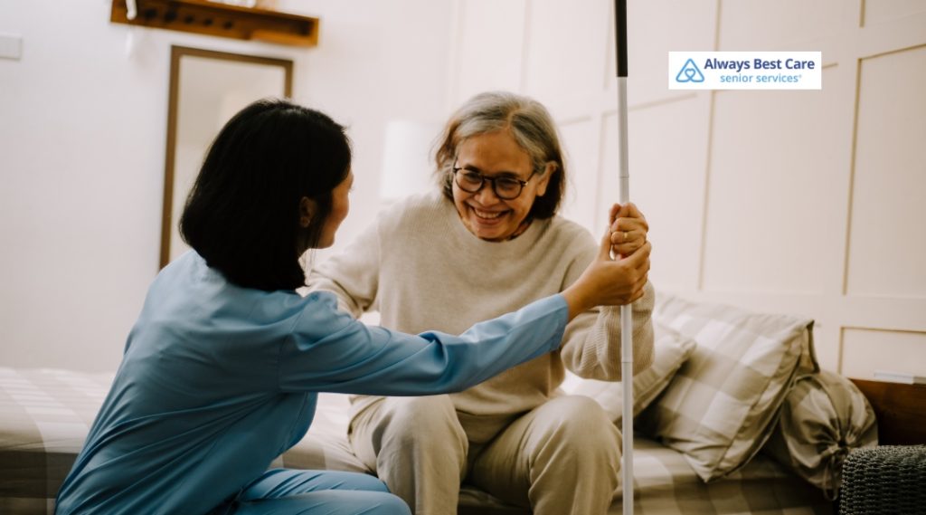 Smiling senior woman sitting on a bed while a caregiver in scrubs offers supportive assistance with a mobility aid in a home setting.