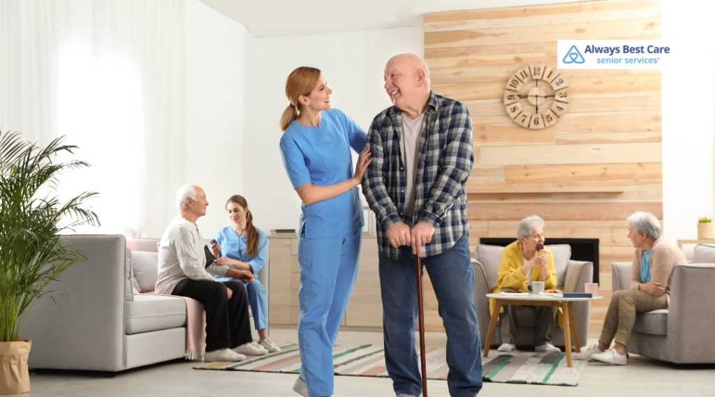 A caregiver assisting a senior man to walk while smiling, with other elderly individuals in the background enjoying a social setting.