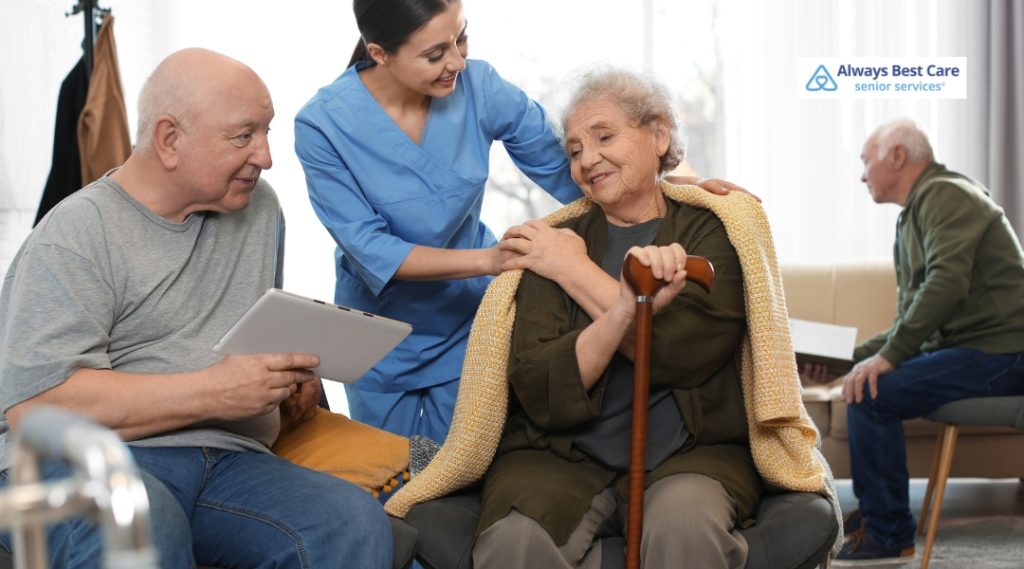 A caregiver helping an elderly woman with a walking cane while engaging with a man, demonstrating supportive in-home care.