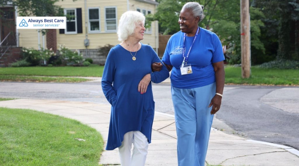 A cheerful caregiver walks arm-in-arm with an elderly woman outside in a residential neighborhood.