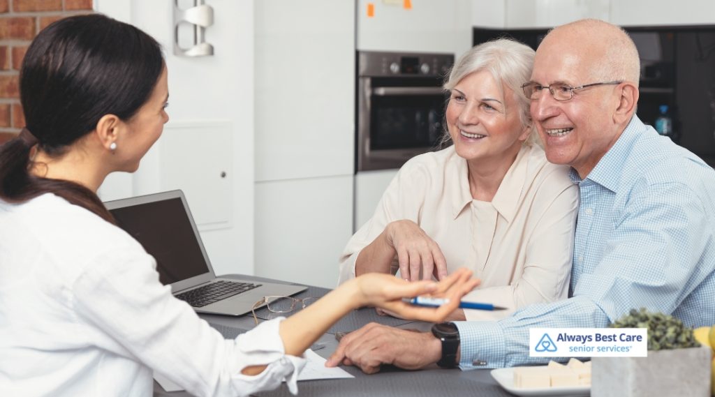 young woman consults with an elderly couple at a kitchen table, referencing paperwork and a laptop. The Always Best Care Senior Services logo appears in the bottom right corner.