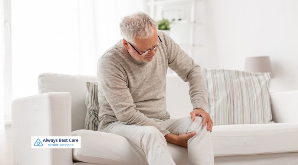  An elderly man sitting on a couch, holding his knee in discomfort, likely experiencing knee pain or discomfort.