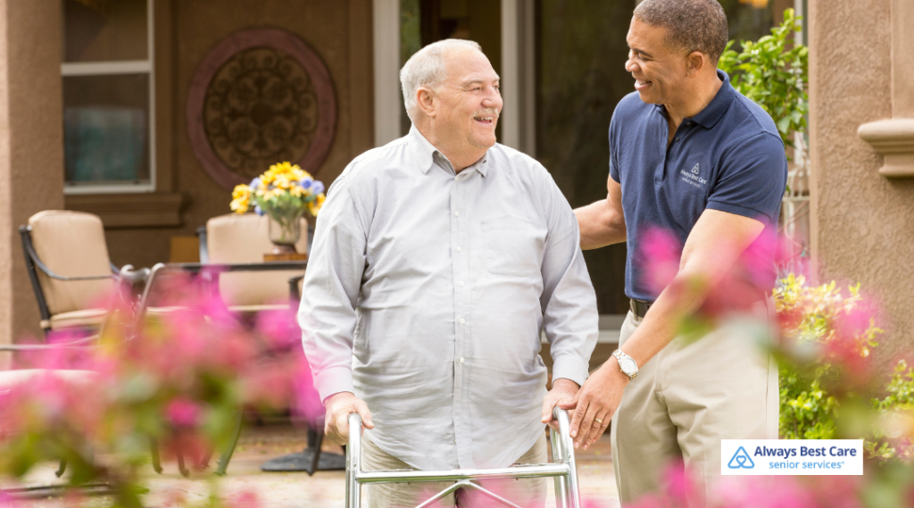 Professional caregiver walking alongside an older man using a walker, offering steady support for safe mobility at home. 
