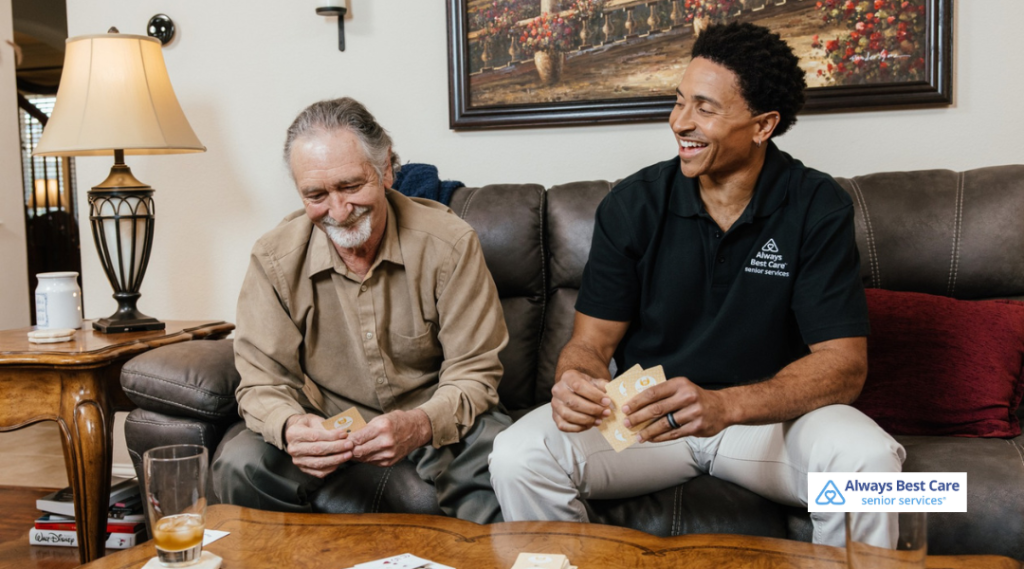 Senior man and caregiver smiling while playing a card game together on a living room couch.
