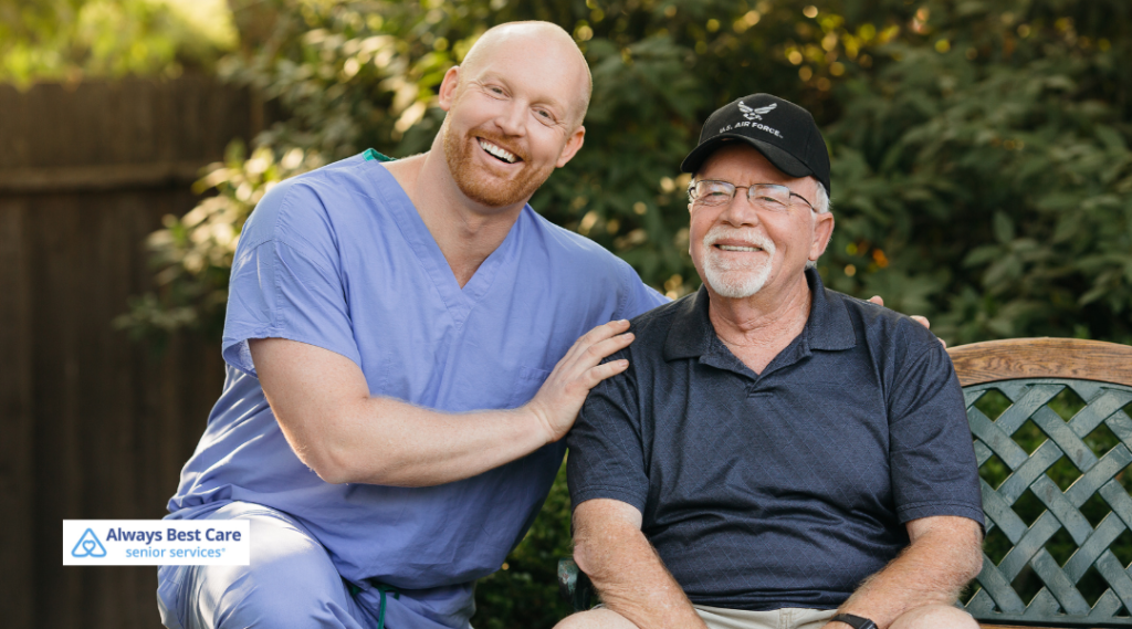 A professional caregiver in light blue scrubs and an older man wearing a black "U.S. Air Force" hat and glasses sit together on a wooden garden bench, both smiling warmly, representing friendly support and companionship for veterans.